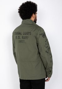 Grøn militær-inspireret jakke lavet af slidstærkt stof. Har tekst på ryggen: "SIGNAL CORPS U.S. NAVY" og grafiske detaljer på ærmerne.
