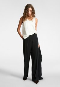 Top bianco senza maniche abbinato a pantaloni neri larghi a vita alta. La modella indossa scarpe marroni a punta, mostrando una semplice collana in oro.