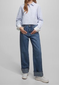Donna che indossa una camicia corta a righe blu, jeans a vita alta con gambe larghe e ampi risvolti, e sneakers bianche, in piedi con le mani in tasca.