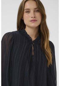 Giovane donna con lunghi capelli castano chiaro che indossa una camicetta nera trasparente plissettata con colletto arricciato e fiocco al collo.