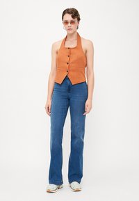Oranža rieva halter top ar sešām pogām un divām priekšējām kabatām, pārī ar ziliem platām biksēm un gaišām kedām.