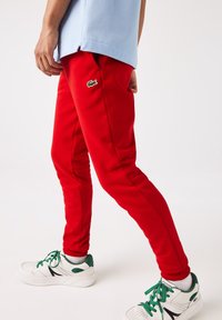 Persoon in rode sweatpants met een groen krokodillenlogo, witte sneakers met groene veters en een lichtblauw shirt, staand tegen een witte achtergrond.