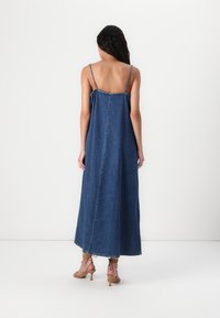 Lang denimkjole med tynne spaghetti-stropper, en rett silhuett og frynsete kant. Har en ensfarget blå farge og subtile sømdetaljer.