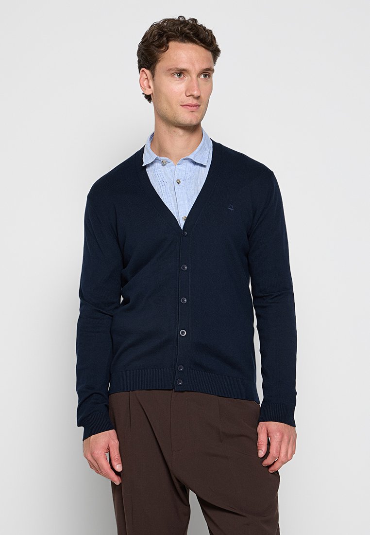 Uomo che indossa un cardigan blu navy a bottoni sopra una camicia colletto azzurra e pantaloni marroni, in piedi contro uno sfondo grigio chiaro uniforme.