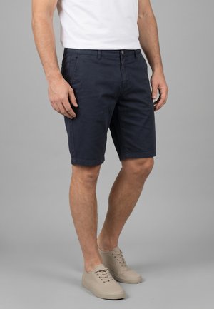 Man draagt marineblauwe knielange shorts, wit T-shirt en beige sneakers staand tegen een effen grijze achtergrond.