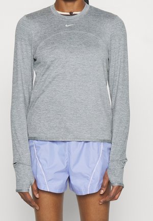 Personne portant un haut de sport gris à manches longues avec des trous pour les pouces et un short de course violet clair, debout devant un fond uni.