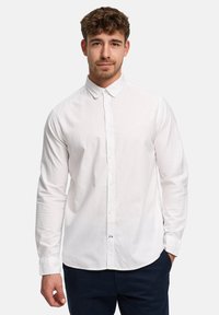 Homme aux cheveux courts et bouclés, portant une chemise blanche à manches longues boutonnée et un pantalon sombre, debout avec une main dans la poche.
