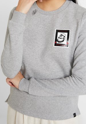 Person trägt ein hellgraues Langarmshirt mit einem Aufnäher eines Cartoon-Matrosen und einer kleinen, bestickten Biene nahe am Kragen.