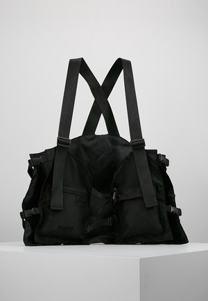 Czarna taktyczna kamizelka z regulowanymi paskami na ramiona, licznymi przednimi kieszeniami i bocznymi klamrami, prezentowana na białym podeście na jednolitym tle.