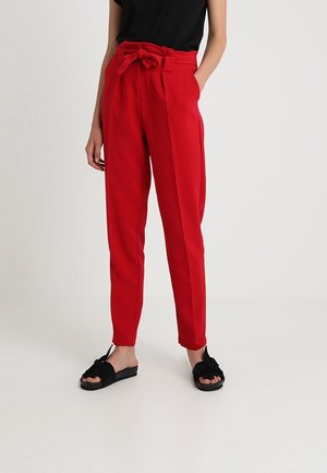 Femme portant un pantalon paperbag rouge taille haute avec une ceinture nouée, un haut noir sans manches et des sandales slides noires avec des nœuds.