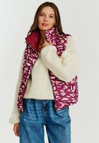 Překřížená vesta v purpurové barvě s růžovým abstraktním vzorem, s vysokým límcem, zipovým zapínáním a hladkým povrchem, nošená přes chlupatý bílý svetr.