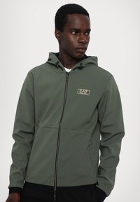 Felpa verde con zip realizzata in tessuto liscio e elasticizzato. Presenta una zip frontale, polsini elasticizzati e una piccola patch con logo sul petto.