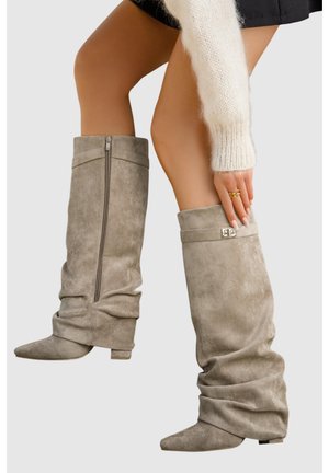 Bottes grises en suède souples montantes jusqu'aux genoux, avec bouts pointus et fermeture éclair latérale, portées avec une jupe noire et un pull blanc en matière duveteuse.