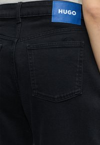 Close-up af baglommen på sorte jeans med en blå rektangulær label med teksten "HUGO" nær linningen og en synlig arm på højre side.