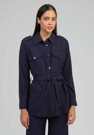 Femme aux longs cheveux foncés portant une veste bleu marine ceinturée avec des boutons et deux poches poitrine, debout devant un fond gris uni.