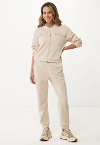 Béžová souprava domácího oblečení obsahuje zipovou bundu se dvěma náprsními kapsami a odpovídající kalhoty s elastickými manžetami. Materiál se zdá být hladký a jemný.