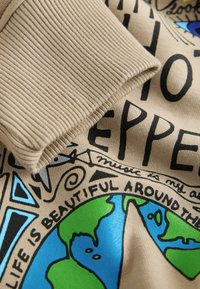 Beige sweatshirt med ribbede manchetter, flerfarvet grafik af Jorden og sorte tekstaccenter. Stoffet virker blødt og struktureret.