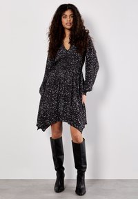Schwarz-weiß gemustertes Kleid mit tiefem V-Ausschnitt, gerafftem Oberteil, langen Ballonärmeln und ungleichem Saum. Kombiniert mit knielangen schwarzen Stiefeln.