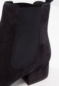 Botte Chelsea en daim noir avec panneau élastique latéral et talon bloc bas sur fond blanc.