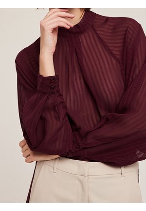 Person iført en langærmet, halv-gennemsigtig bordeauxbluse med lodrette striber og høj hals, kombineret med lyse beige bukser.