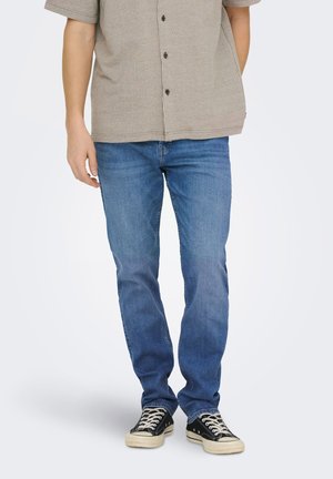 Homme portant un jean bleu, des baskets noires et blanches, et une chemise beige à manches courtes boutonnée, debout devant un fond uni.