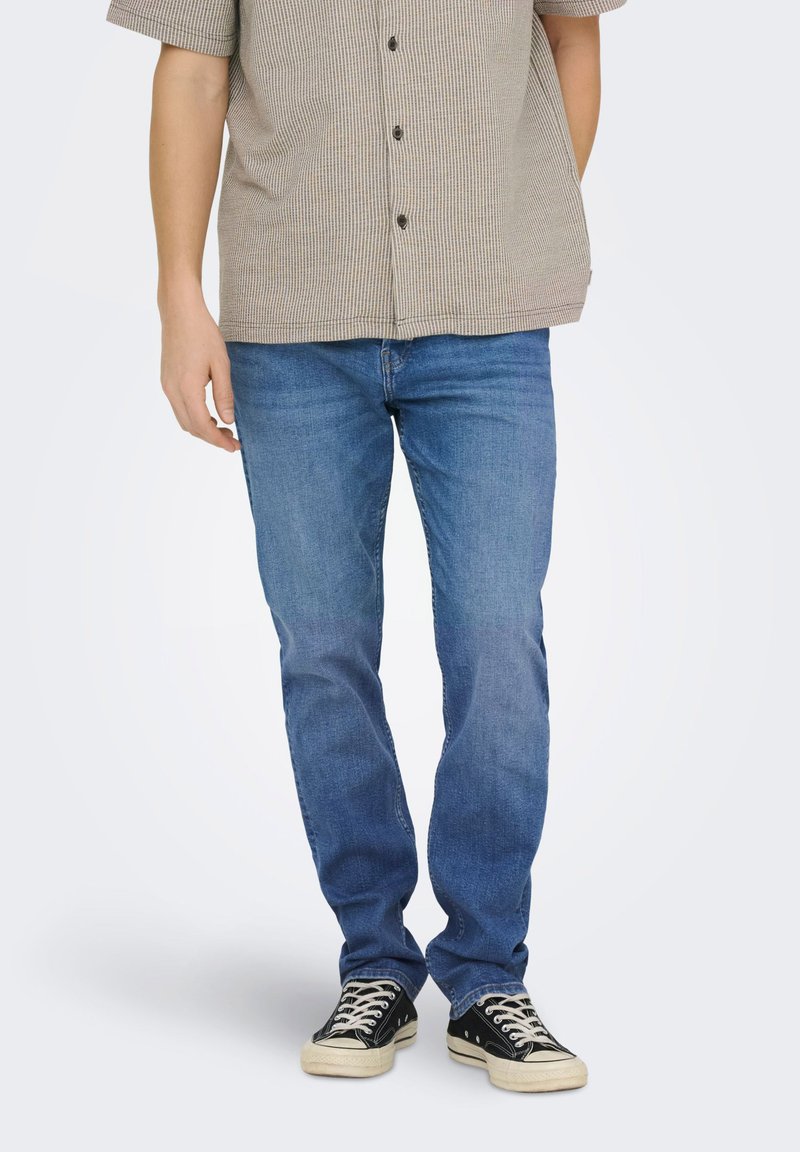 Homme portant un jean bleu, des baskets noires et blanches, et une chemise beige à manches courtes boutonnée, debout devant un fond uni.