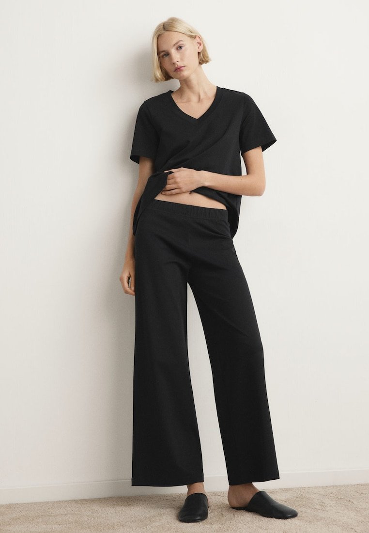 Femme blonde en haut noir à manches courtes et col en V, portant un pantalon noir à jambes larges, pieds nus sur un tapis contre un mur blanc.