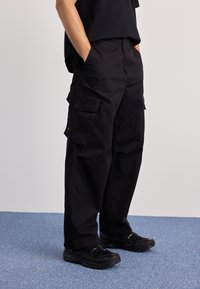 Schwarze Cargo-Hose mit lockerer Passform, die mehrere Seitentaschen, eine Gesäßtasche und einen Knopfverschluss hat. Tragen mit schwarzen Sneakers.