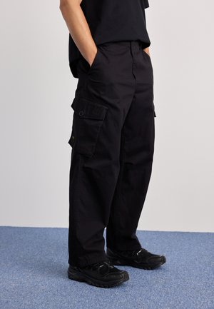 Schwarze Cargo-Hose mit lockerer Passform, die mehrere Seitentaschen, eine Gesäßtasche und einen Knopfverschluss hat. Tragen mit schwarzen Sneakers.