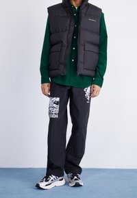 Gilet imbottito nero sopra una camicia verde con bottoni, abbinato a pantaloni neri con una toppa grafica bianca. Indossato con sneaker argentate.