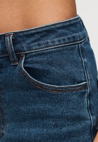 Κοντινό πλάνο της μέσης μπλε τζιν με πορτοκαλί ραφές και εμφανή θηλιά ζώνης πάνω σε φόντο δέρματος.