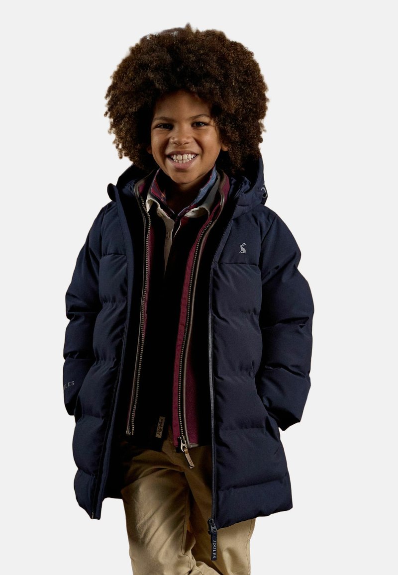 Enfant souriant aux cheveux bouclés portant une doudoune bleu marine, des chemises superposées et un pantalon beige sur un fond uni.