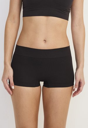 Frau trägt schwarze nahtlose Boyshort-Unterwäsche und ein schwarzes Crop-Top, stehend vor einem einfarbigen hellen Hintergrund.