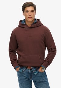 Felpa con cappuccio color bordeaux, dotata di tasca frontale e un piccolo logo. Abbinata a una camicia a quadri e jeans blu, presenta un look casual sovrapposto.