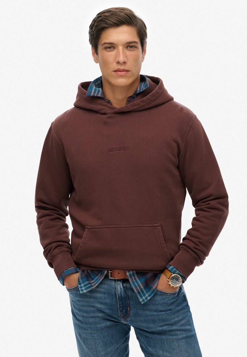 Felpa con cappuccio color bordeaux, dotata di tasca frontale e un piccolo logo. Abbinata a una camicia a quadri e jeans blu, presenta un look casual sovrapposto.