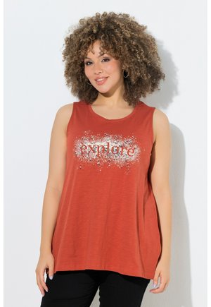 Frau mit lockigem Haar, die ein ärmelloses Top in Rostfarbe mit dem Wort „explore“ im weißen Spritzdesign trägt und vor einem einfarbigen Hintergrund lächelt.