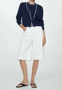 Cardigan marine avec passepoil blanc, fermeture à boutons et manches longues, associé à un short en denim blanc Bermuda avec ourlet effiloché, porté avec des tongs noires.