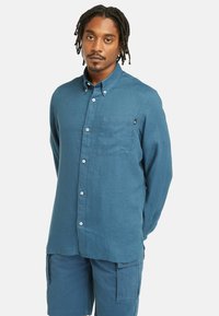 Camisa de manga comprida em azul, feita de tecido texturizado. Apresenta um colarinho abotoado, bolso frontal e botões brancos. Combinada com calções cargo azuis.