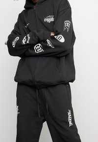 Ensemble sweat à capuche zippé noir et pantalon de survêtement, avec plusieurs logos et graphiques blancs sur les bras et les jambes. Tissu doux avec une coupe décontractée.