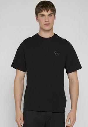 Ung mand iført en ensfarvet sort kortærmet T-shirt med lille sort trekantet logo på venstre bryst, stående foran en lys grå baggrund.