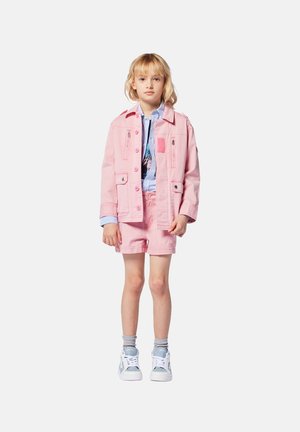 Veste en denim rose avec poches avant, assortie à un short rose. En dessous, une chemise rayée bleue et blanche. Baskets blanches avec des accents gris.
