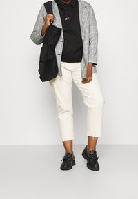 Blazer de cuadros gris sobre una sudadera con capucha negra, pantalones recortados crema y zapatos negros con un lazo. Una bolsa tote negra está colgada del hombro.