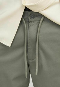 Los pantalones verde oliva tienen cordones y cierre de botón. Textura de tela suave con un corte moderno. La capa superior muestra un cuello de camisa color crema.