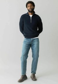 Marineblauer Rippensweater mit halb geöffnetem Reißverschluss, kombiniert mit hellblauen Jeans und braunen Schnürstiefeln. Das Modell steht mit gefalteten Händen an der Taille.