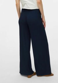 Weite Hose aus strukturiertem, dunkelblauem Material mit hohem Bund. Glatte Oberfläche ohne sichtbare Akzente. Getragen mit einem hellen Oberteil und braunen Schuhen.