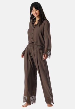 Kvinde smilende og stående barfodet iført løstsiddende brune pyjamas med blonder på ærmer og ben mod en ensfarvet baggrund.