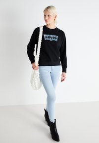 Melnā sporta krekls ar košām "Levi's" grafikām, gaiši zilas šaurās džinsas, teksturēta balta soma un melni potīšu zābaki. Vienkāršs dizains, ikdienišķs piegriezums.