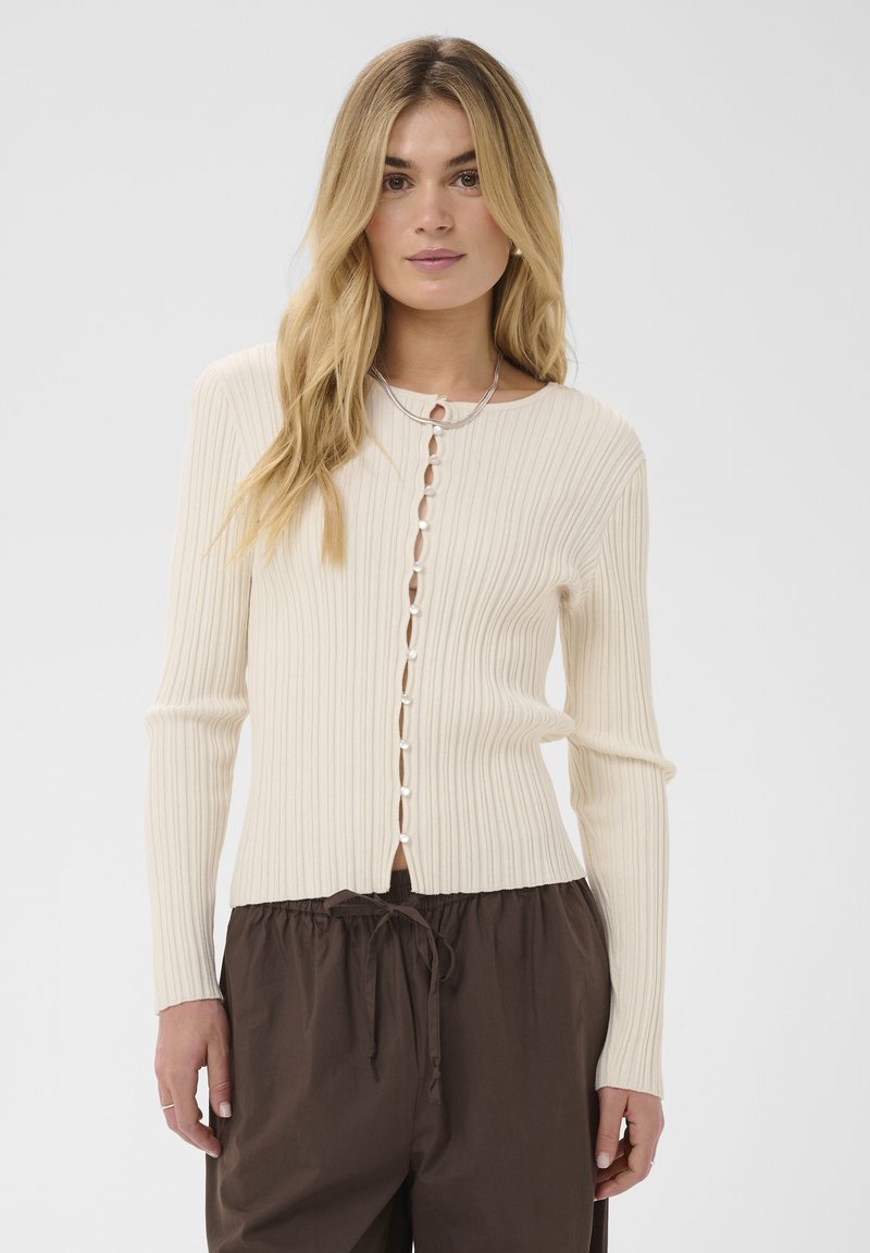 Femme portant un cardigan côtelé crème avec des boutons en perle et un pantalon marron foncé à cordon, debout devant un fond clair uni.