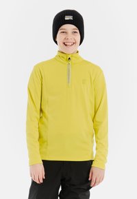 Limoengroene langemouwen top met een halve rits, logo op de borst, gecombineerd met zwarte broeken. Ggedragen met een zwarte geribde beanie met een patch.