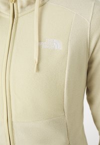Chaqueta de forro polar en beige suave, con cremallera frontal, dos bolsillos laterales y un logo bordado en el pecho. Presente detalle de tejido texturizado.